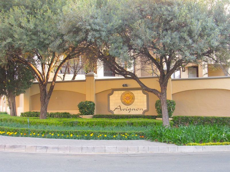 The front entrance of the Avignon apartment complex, featuring a decorative boundary wall, manicured garden beds, and mature trees.