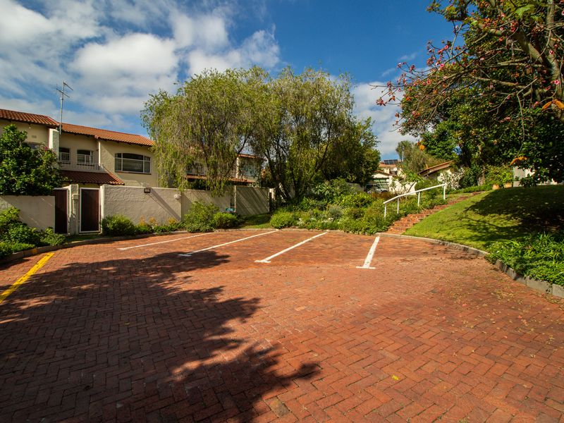 A well-maintained exterior view of the townhouse complex featuring a brick-paved parking area, landscaped garden, and established trees.