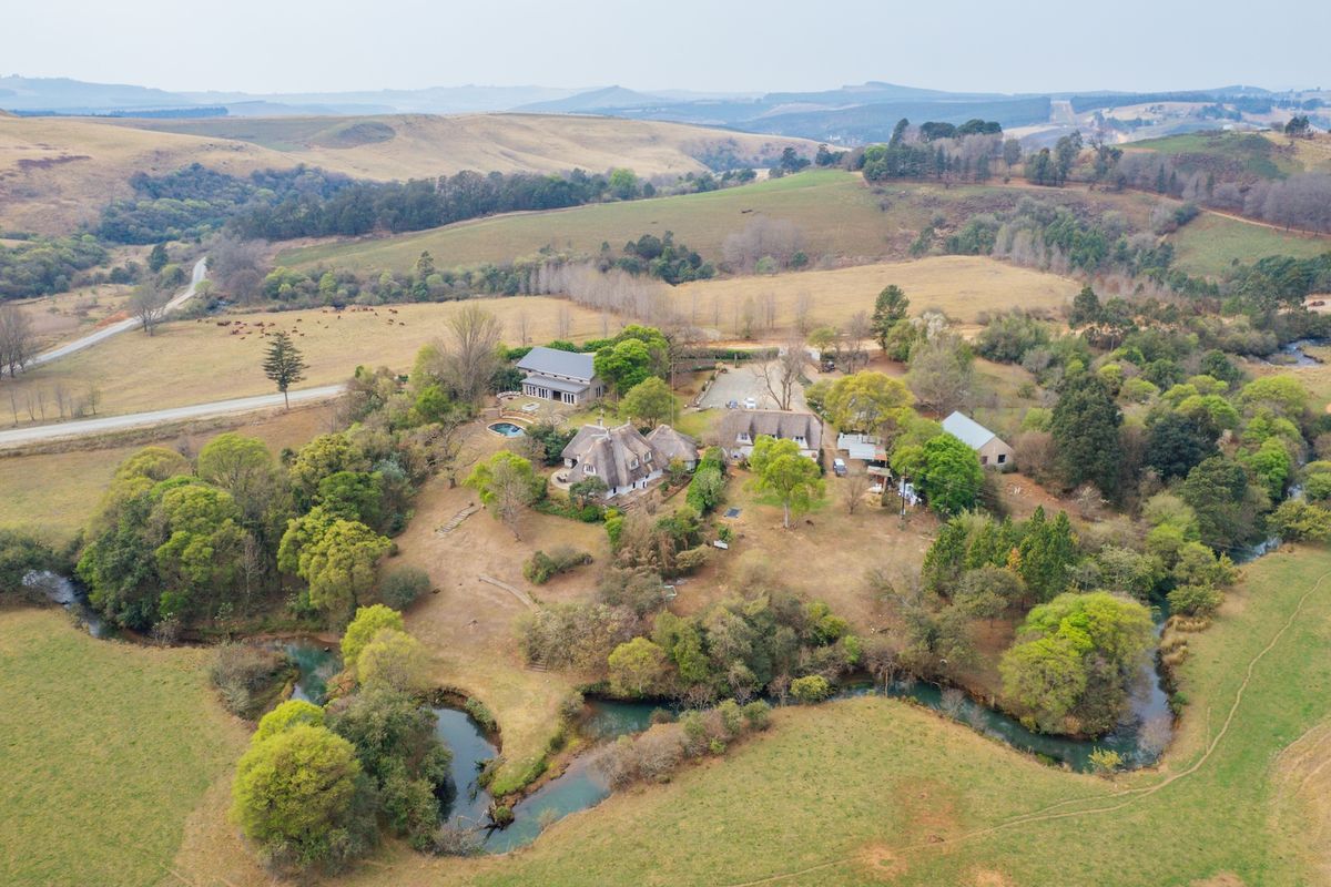Aerial view of a sprawling small holding featuring multiple thatched-roof buildings, a swimming pool, and extensive grounds with river frontage in a rural landscape.