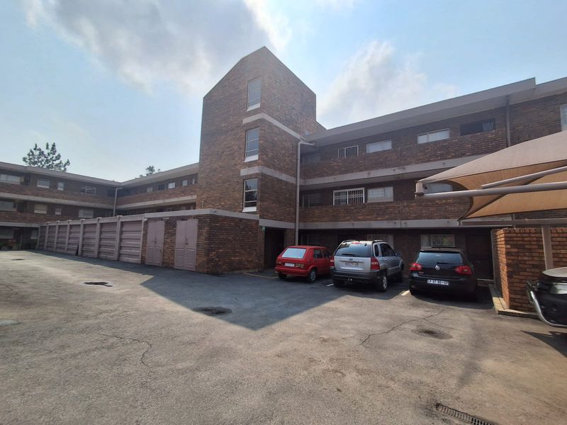 Exterior view of a multi-story face-brick apartment complex featuring a paved parking area, garage units, and a shade sail structure.