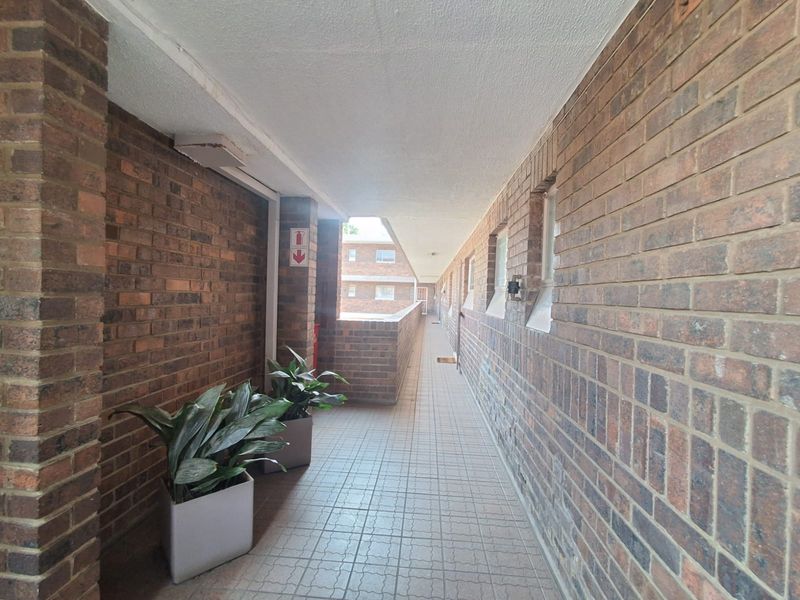 An open-air apartment complex hallway featuring exposed brick walls and tiled flooring, providing access to multiple residential units.