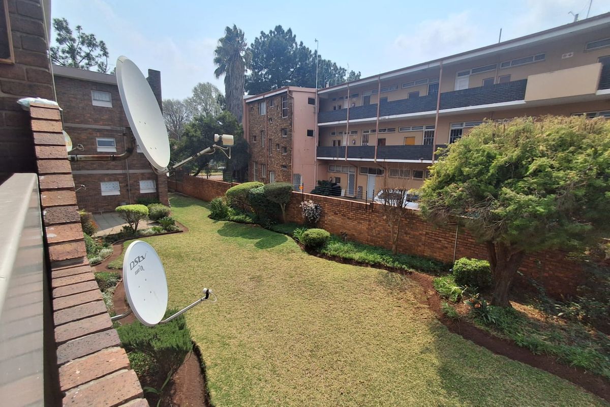 A view of the communal rear garden area featuring a well-maintained lawn, mature trees, and brick boundary walling within the flat complex.