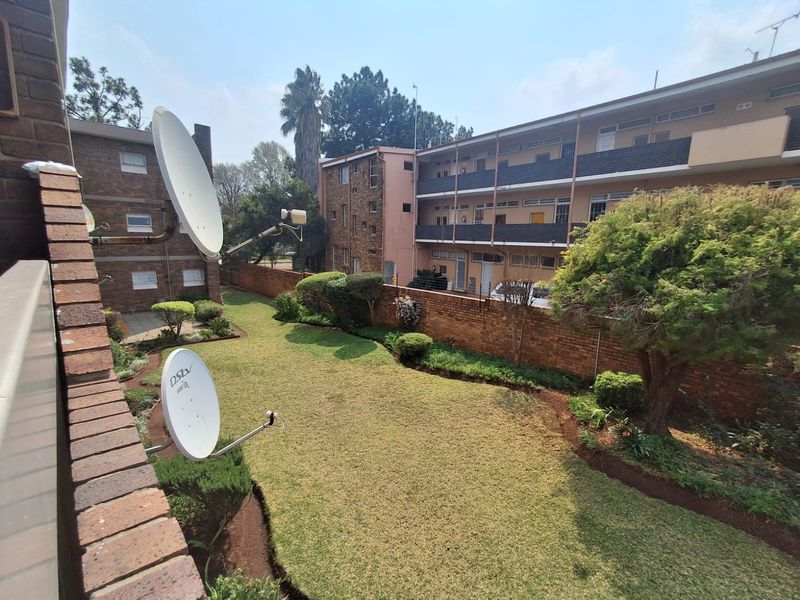 A view of the communal rear garden area featuring a well-maintained lawn, mature trees, and brick boundary walling within the flat complex.