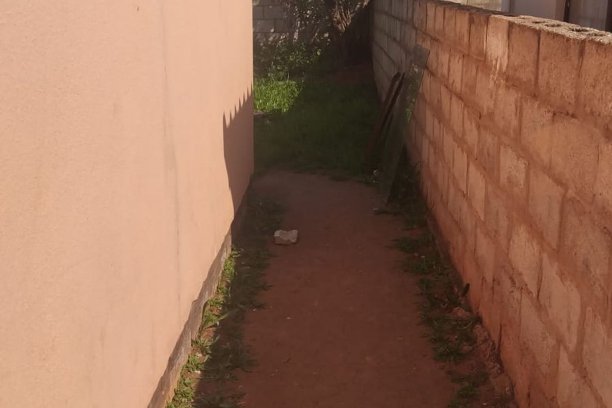 A narrow side passage between the house exterior and a concrete block boundary wall, featuring an unpaved earth pathway.