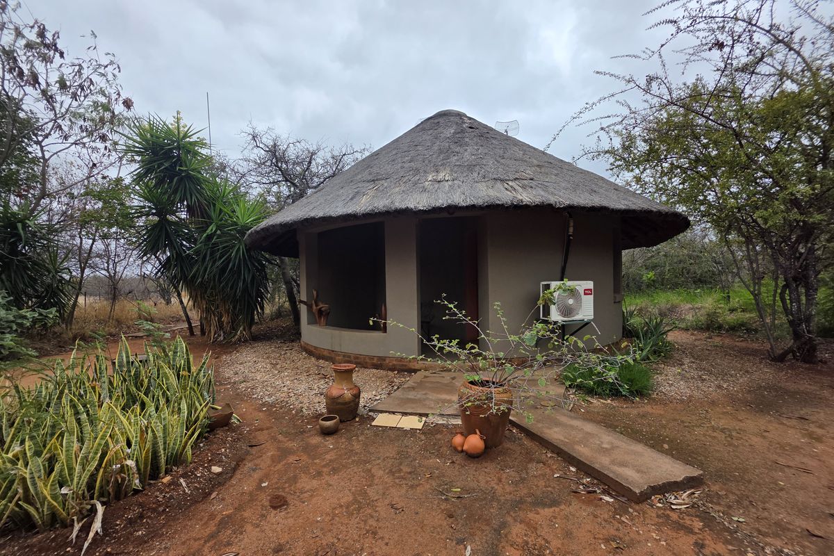 A rustic round cottage with a traditional thatched roof, featuring an exterior air conditioning unit and a concrete pathway set within a natural garden.