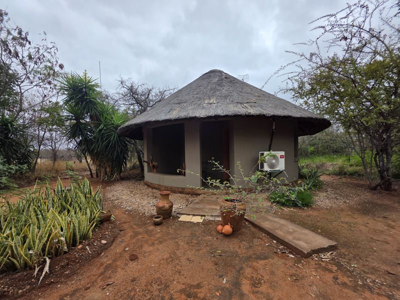 A rustic round cottage with a traditional thatched roof, featuring an exterior air conditioning unit and a concrete pathway set within a natural garden.