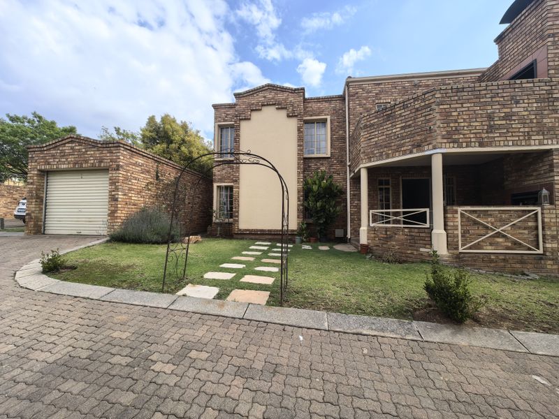 Front elevation of a face brick townhouse featuring a single garage, paved driveway, and a covered patio entrance with a landscaped garden.