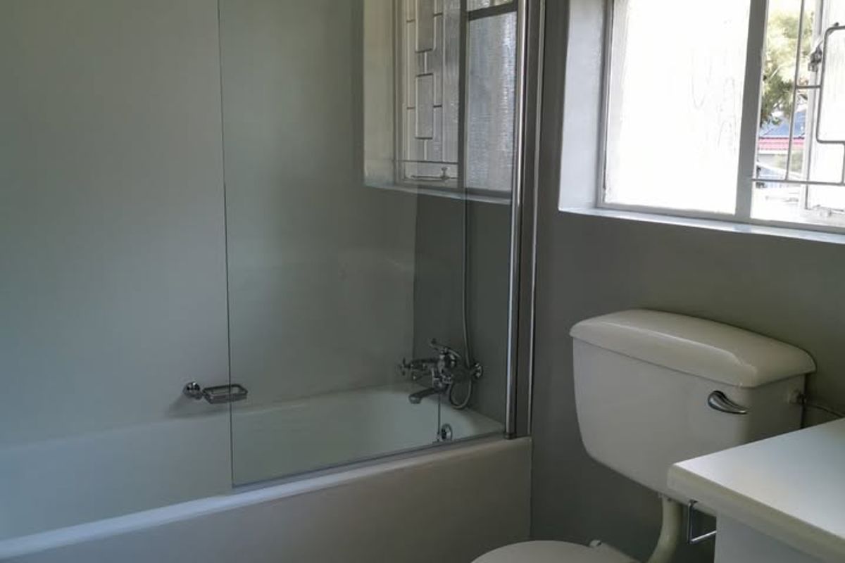 A functional bathroom featuring a bathtub with a glass shower screen, a standard toilet, and a vanity cabinet under a window with security bars.