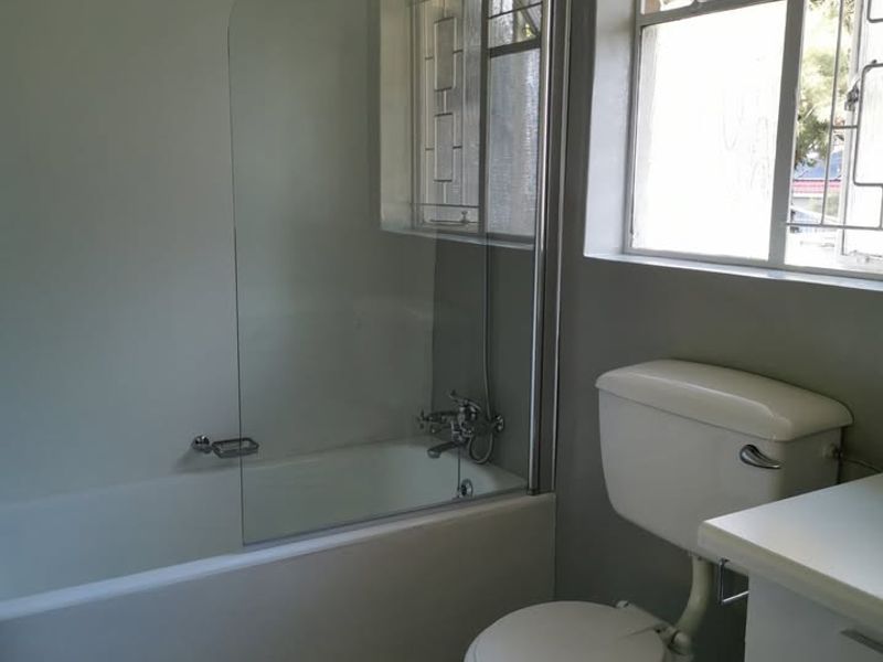 A functional bathroom featuring a bathtub with a glass shower screen, a standard toilet, and a vanity cabinet under a window with security bars.