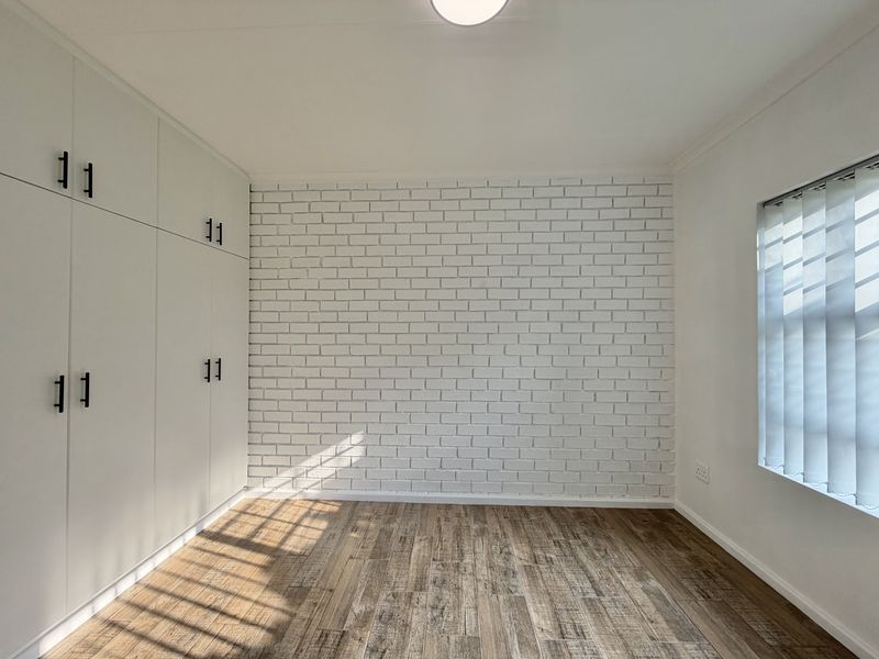 A bright, empty bedroom featuring a white brick accent wall, modern laminate flooring, and extensive built-in cupboard storage.