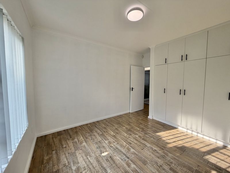 A bright, empty bedroom featuring modern wood-look tiled flooring, ample built-in cupboard storage, and vertical window blinds.