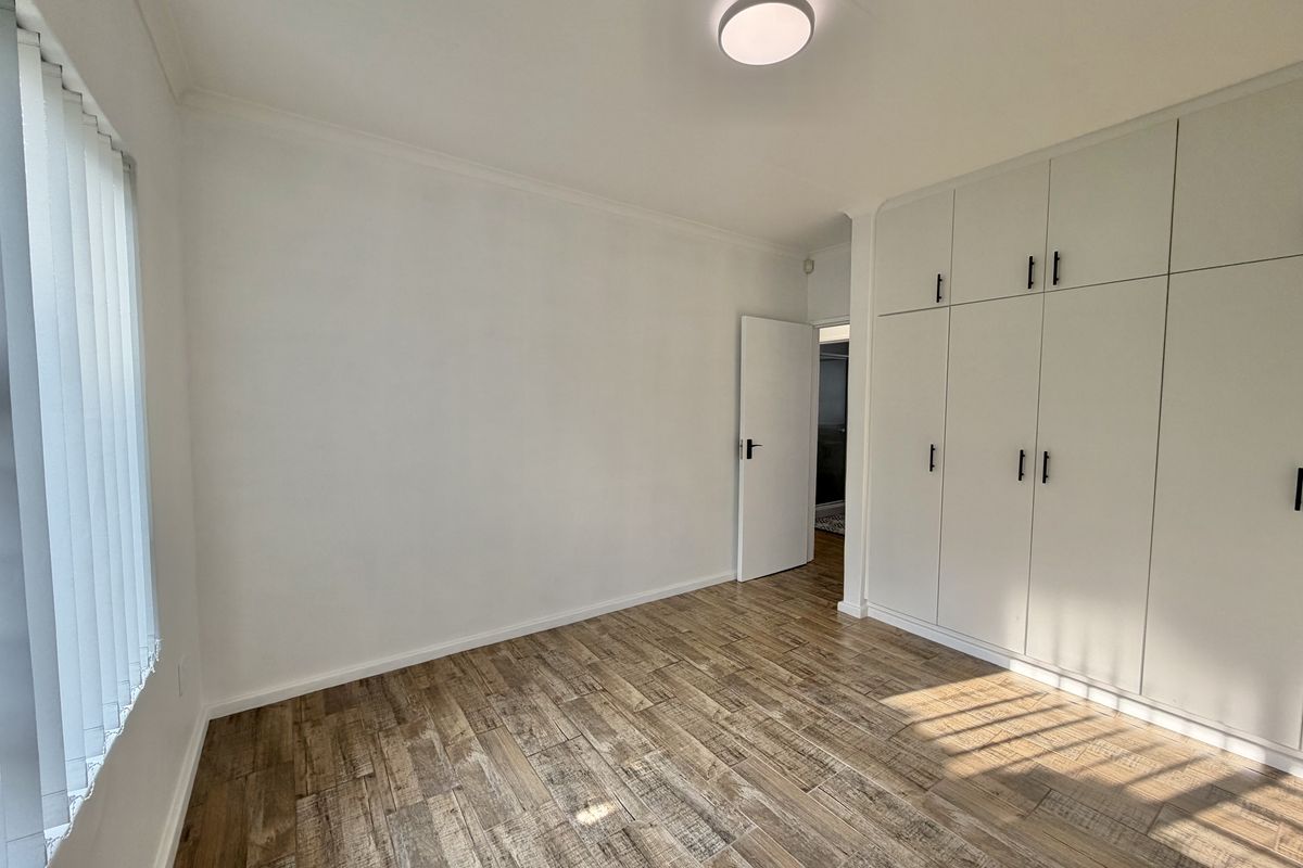 A bright, empty bedroom featuring modern wood-look tiled flooring, ample built-in cupboard storage, and vertical window blinds.
