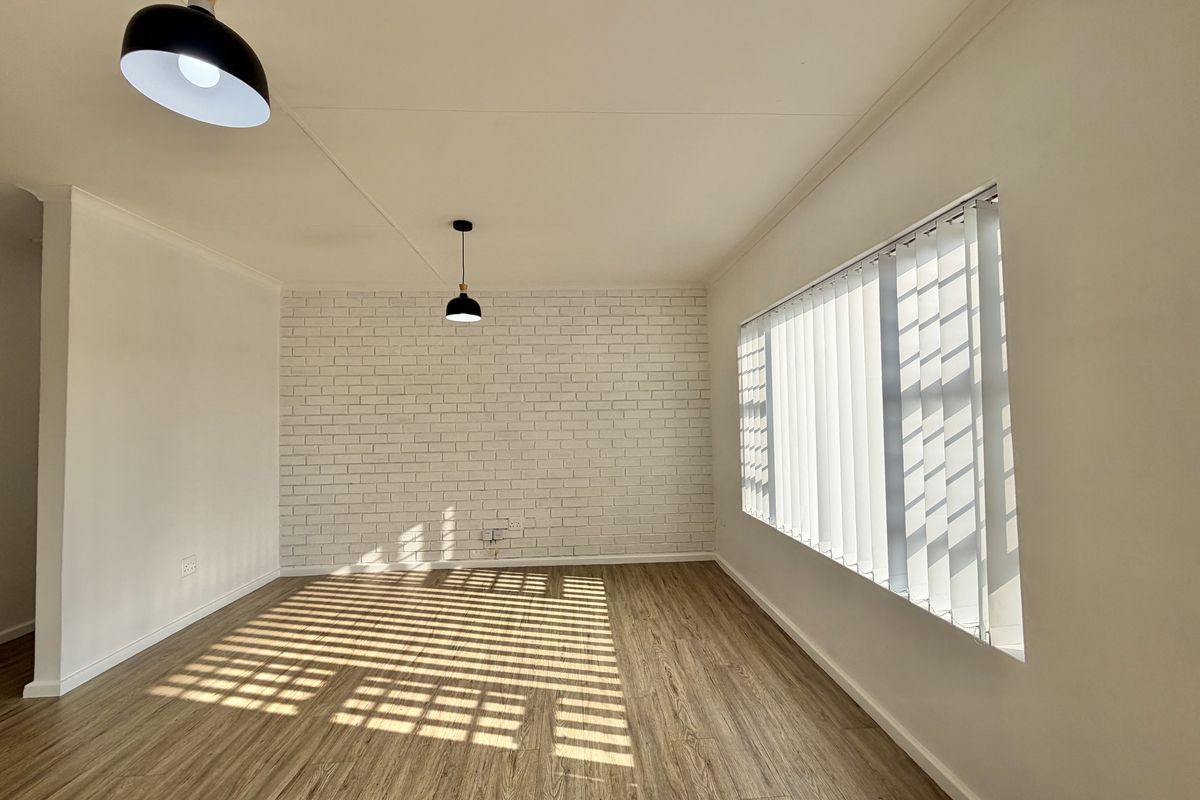 A bright, empty living room featuring modern laminate flooring, a white-painted exposed brick feature wall, and natural light filtered through vertical blinds.
