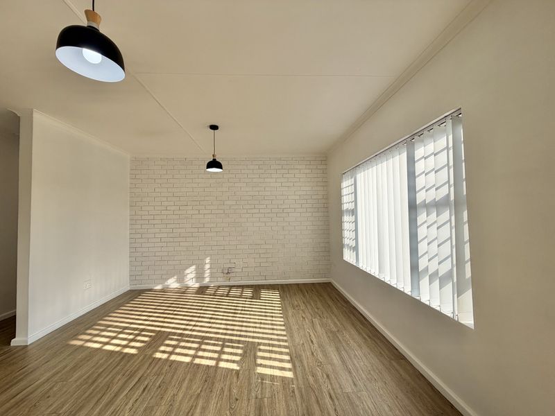 A bright, empty living room featuring modern laminate flooring, a white-painted exposed brick feature wall, and natural light filtered through vertical blinds.