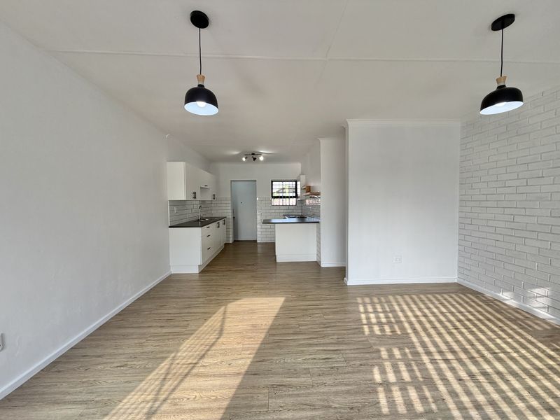 Spacious open-plan living area featuring modern laminate flooring, a white brick accent wall, and a functional kitchen with clean cabinetry and contemporary pendant lighting.