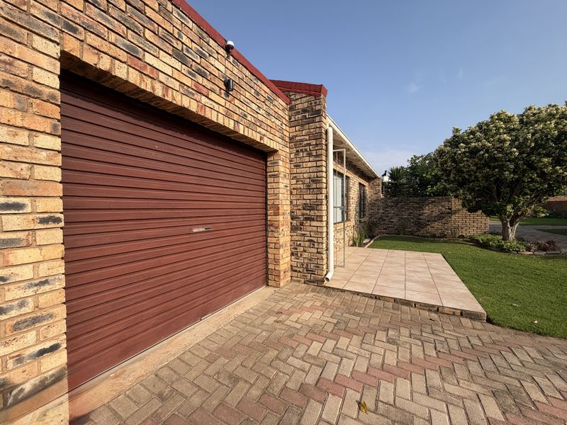 Exterior view of a townhouse featuring a brick facade, a roll-up garage door, a paved driveway, and a small tiled patio area.
