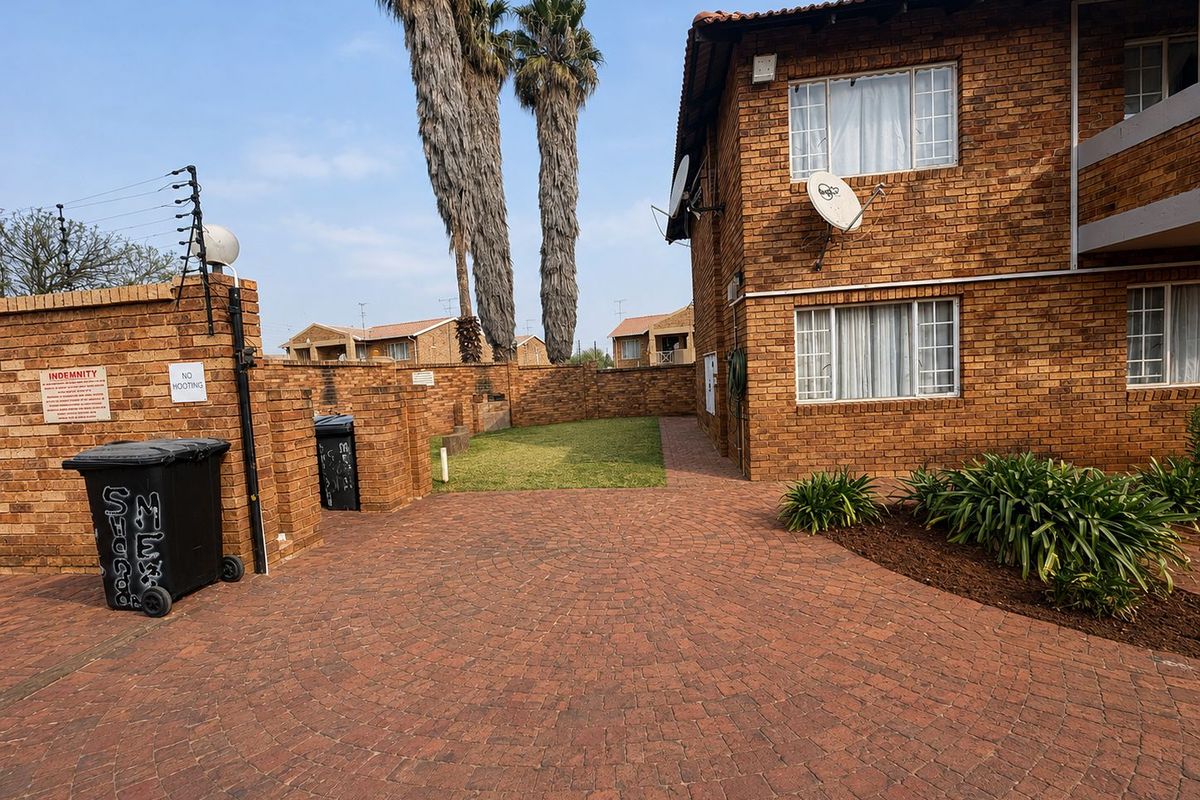 Rear exterior view of the townhouse featuring a brick facade, paved courtyard area, and perimeter security fencing.