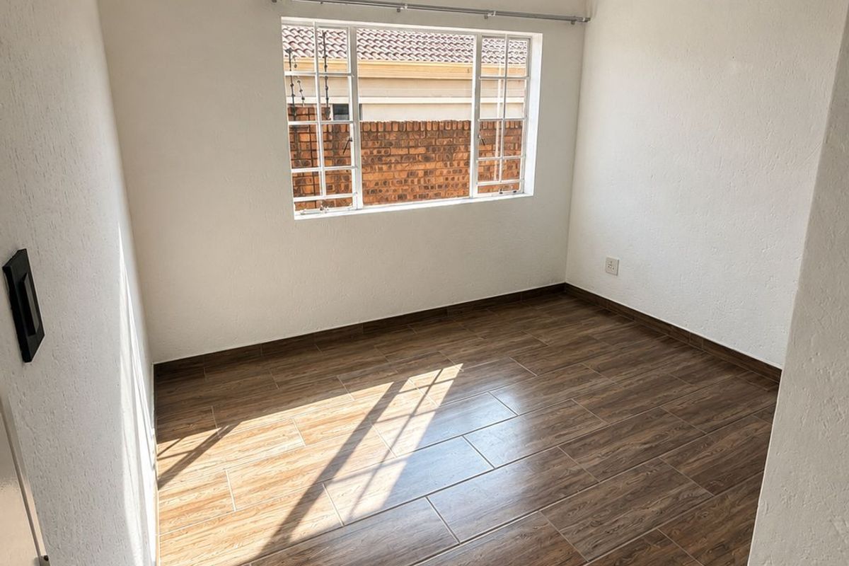 A bright, empty bedroom featuring modern wood-look tiled flooring, a window with security bars, and neutral white walls.