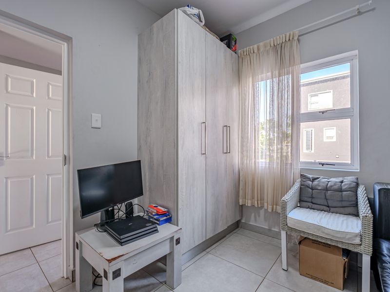 A compact bedroom featuring neutral tiled flooring, a built-in wardrobe with light wood-grain finish, and an aluminium-framed window allowing natural light.
