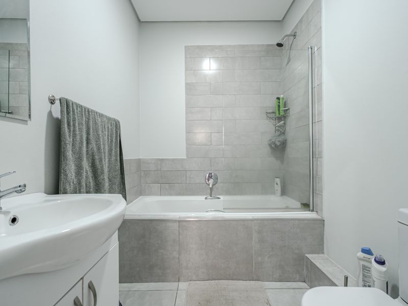 A modern bathroom featuring a vanity basin, a combined bathtub and shower with a glass screen, and neutral grey tiling throughout.