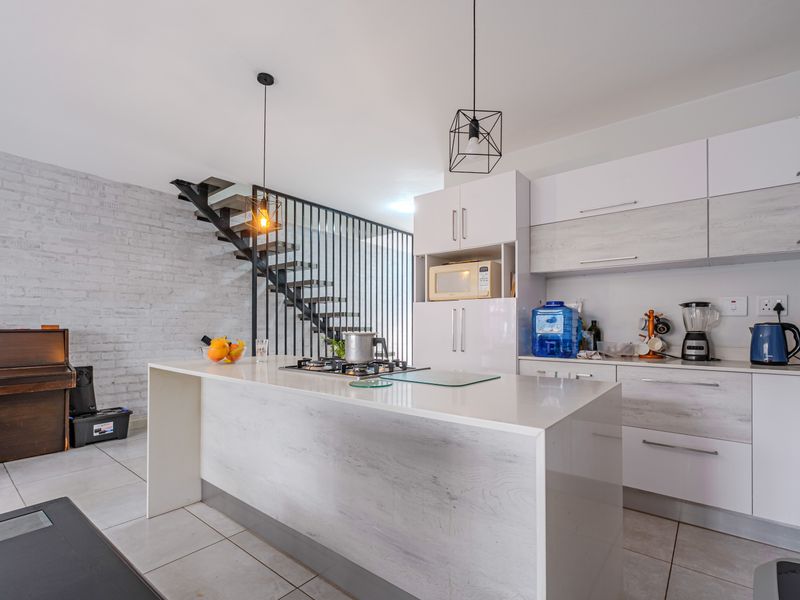 Modern open-plan kitchen featuring a central island, sleek cabinetry, and a striking staircase with a metal balustrade against an exposed brick wall.