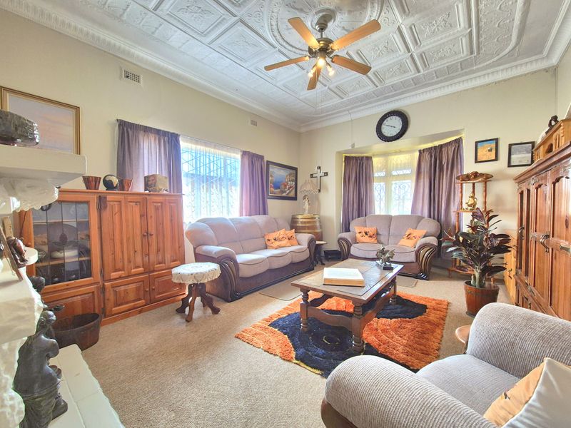 A spacious living room featuring a classic pressed metal ceiling, a ceiling fan, and a bay window with security bars.
