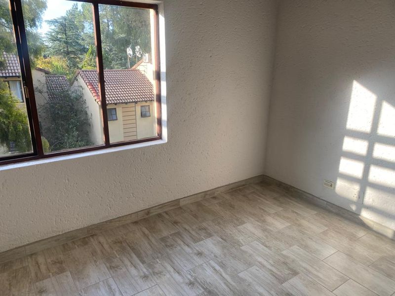 A bright, empty bedroom featuring modern wood-look tiled flooring, textured walls, and a large window with views of the surrounding complex.