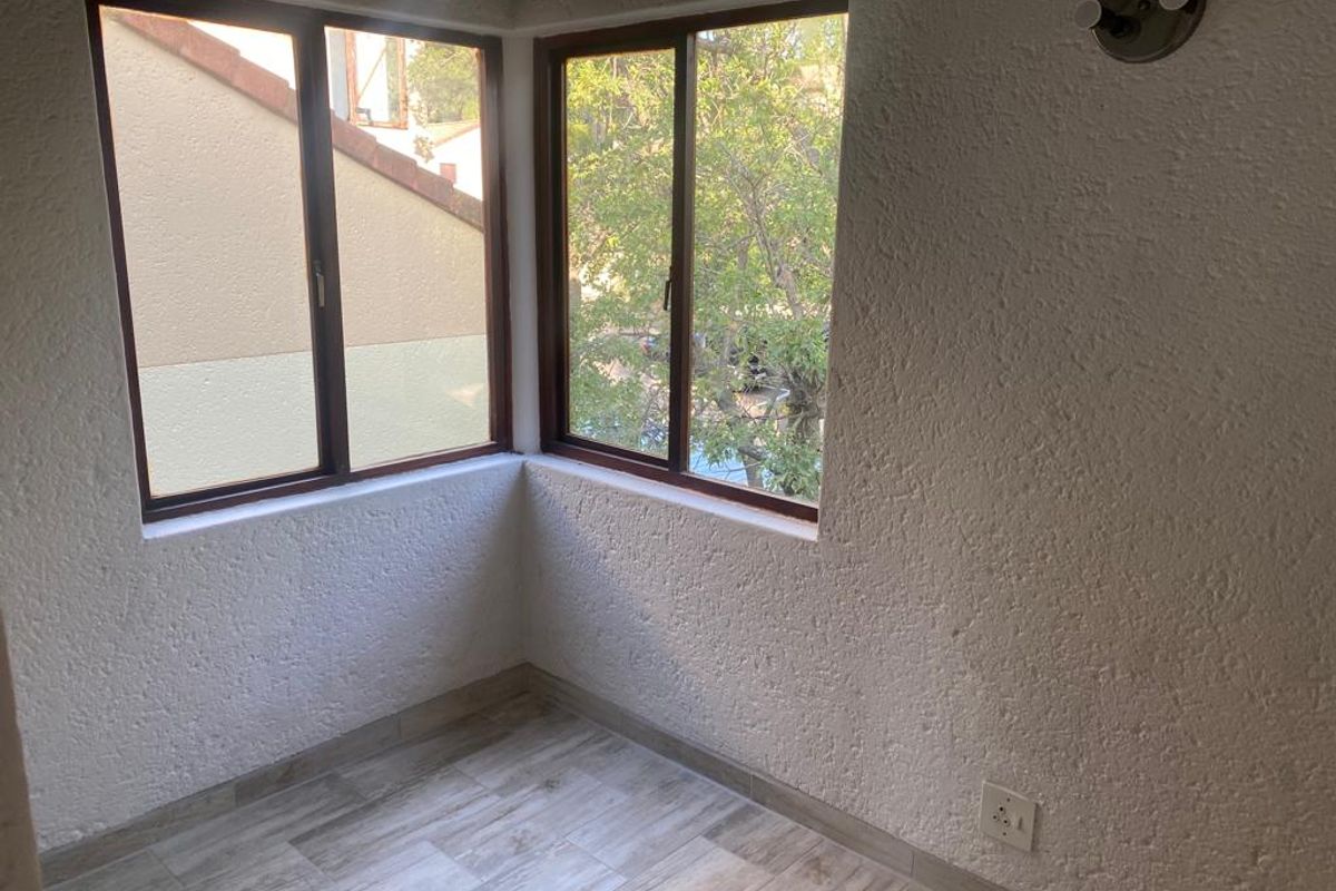 A bright, empty corner room featuring wood-look tile flooring, textured walls, and large windows providing natural light and a view of the surrounding greenery.