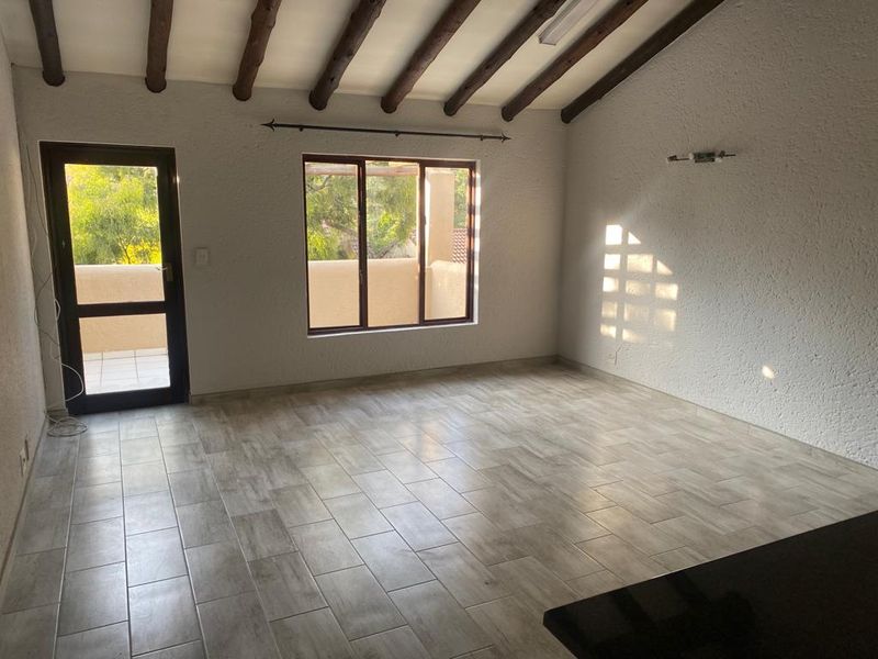 Spacious living area featuring exposed timber ceiling beams, modern tiled flooring, and direct access to an outdoor balcony through a glass panel door.