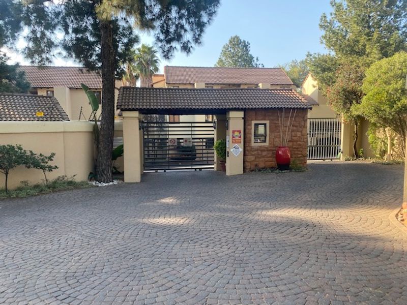 Secure gated entrance to the townhouse complex featuring a cobblestone driveway, stone-clad gatehouse, and perimeter wall with electric fencing.