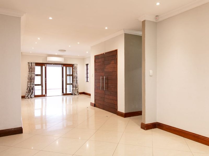 Spacious, open-plan living area featuring neutral tiled flooring, recessed downlighting, and elegant wooden double doors leading to an outdoor space.