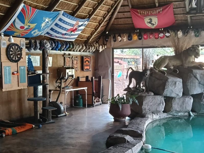 An indoor entertainment area featuring a swimming pool, thatched roof with exposed timber rafters, and large sliding glass doors opening to the garden.