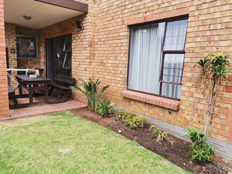 A rear exterior view of the apartment featuring a covered tiled patio, brick facade, and a small private garden area with lawn.