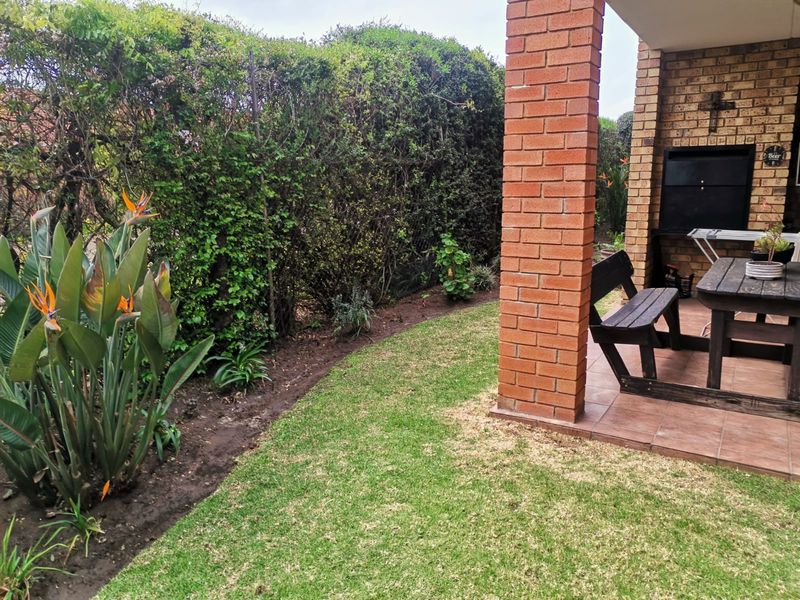 A covered patio area featuring a built-in braai, brick pillar support, and tiled flooring, overlooking a private garden with established hedging.