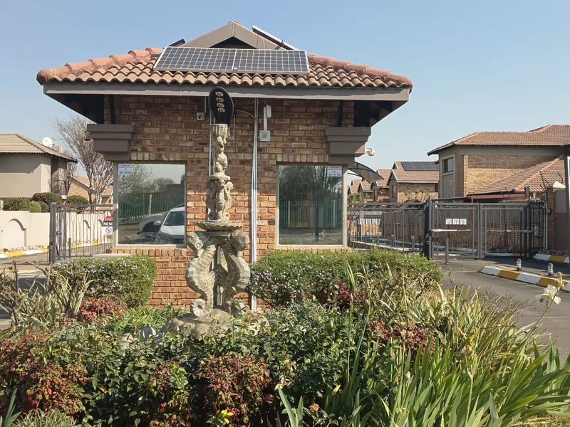 The entrance to the apartment complex features a brick guardhouse with solar panels, secure gated access, and well-maintained landscaped garden beds.