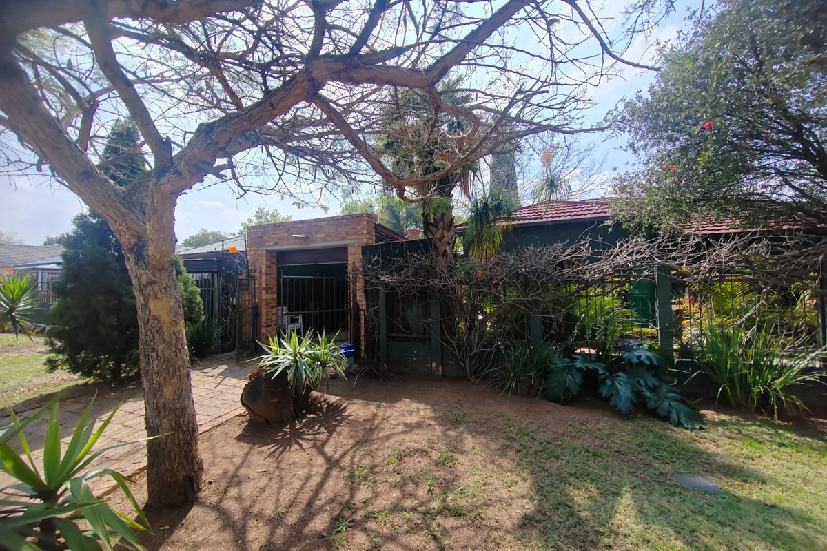 A rear garden view featuring a brick-faced structure, tiled roof, and established trees with a paved walkway leading to a secure gated entrance.