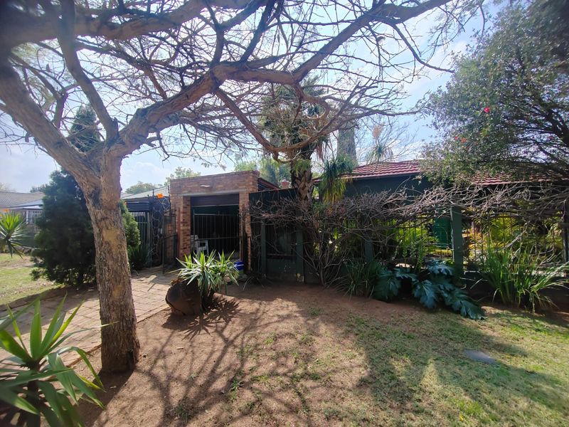 A rear garden view featuring a brick-faced structure, tiled roof, and established trees with a paved walkway leading to a secure gated entrance.