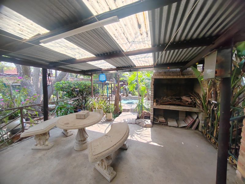 Covered outdoor patio area featuring a built-in braai, concrete flooring, and corrugated roofing, overlooking the garden and pool area.