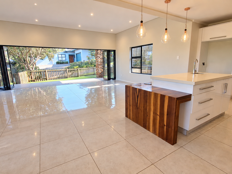 Spacious open-plan living and kitchen area featuring a modern island with stone countertops, wooden breakfast bar, glossy cabinetry, and large bi-fold doors to the exterior.