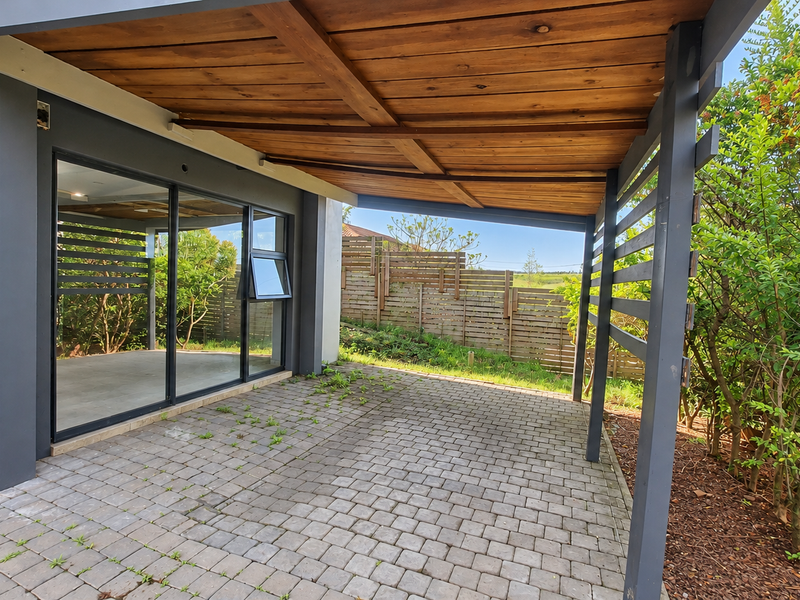 A covered outdoor patio featuring a timber pergola, paved flooring, and sliding glass door access to the interior, bordered by timber slat fencing.