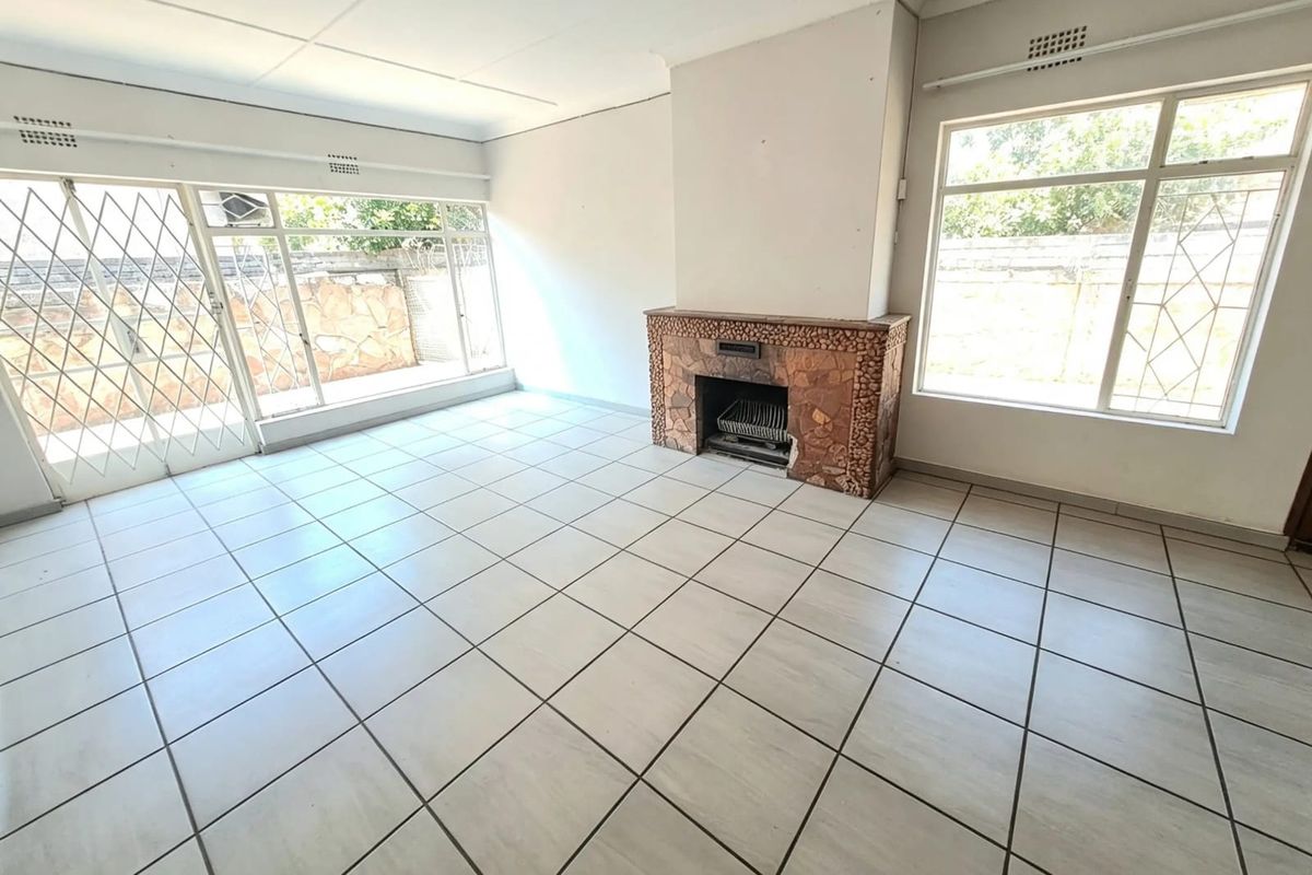 An empty living room featuring tiled flooring, a stone fireplace, and large windows with security gates, offering a blank canvas for renovation.