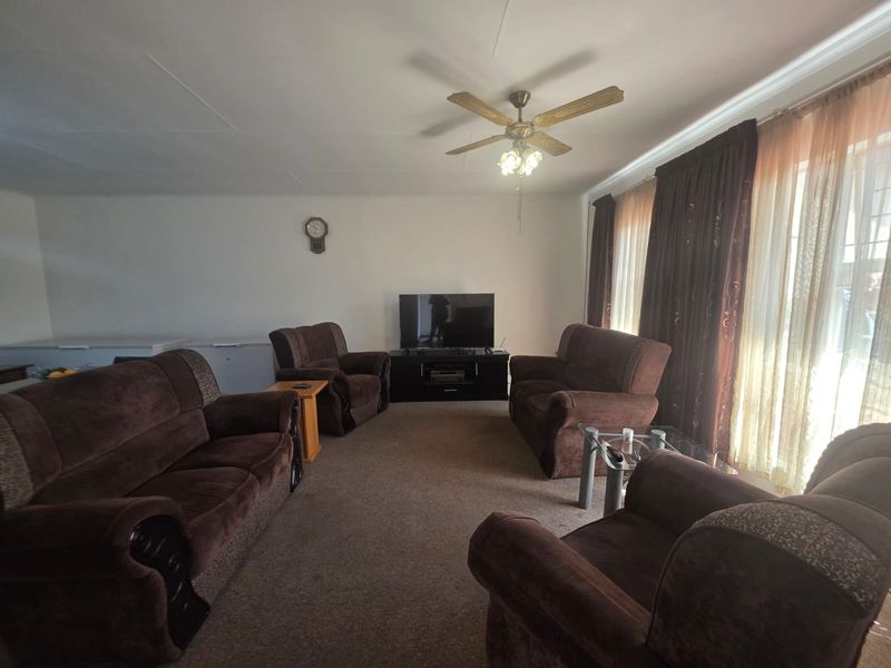A spacious living room featuring carpeted flooring, a central ceiling fan, and large windows with curtain treatments.