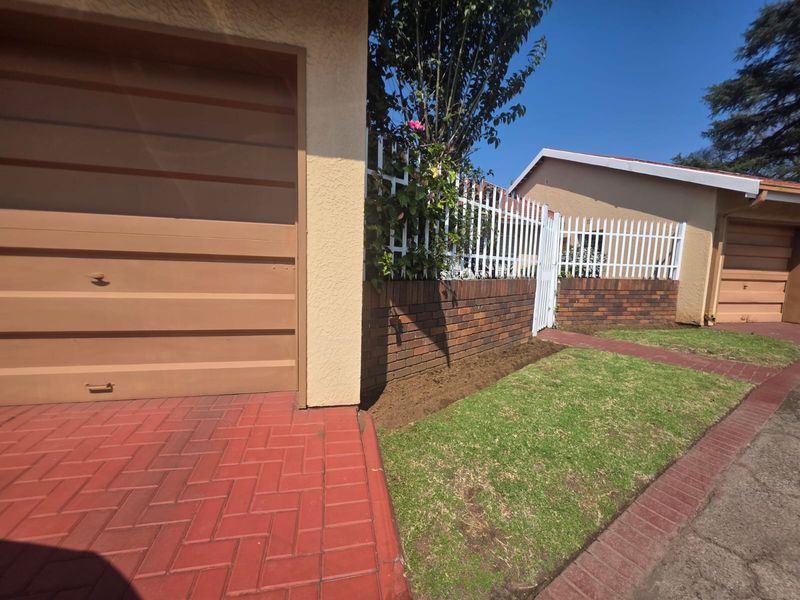 Front exterior view of the townhouse featuring a garage, brick boundary wall with metal fencing, and a paved driveway with a small lawn area.