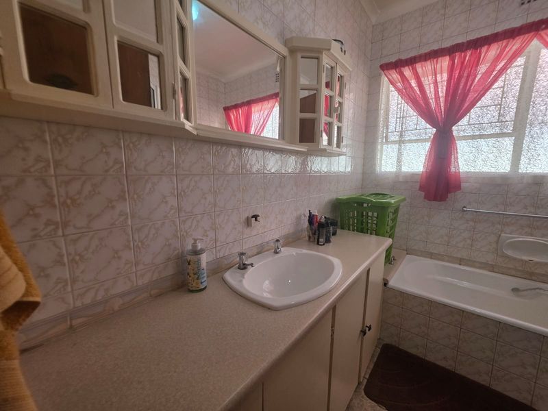 A dated bathroom featuring a built-in vanity with integrated basin, wall-mounted mirror, and a tiled bathtub area with security bars on the window.