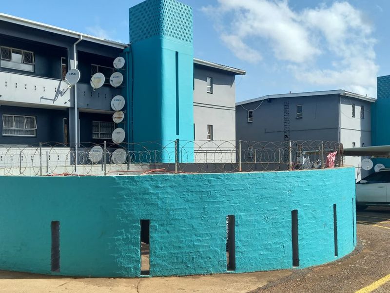 Exterior view of an apartment complex featuring a bright blue boundary wall with razor wire fencing and multiple satellite dishes mounted on the facade.