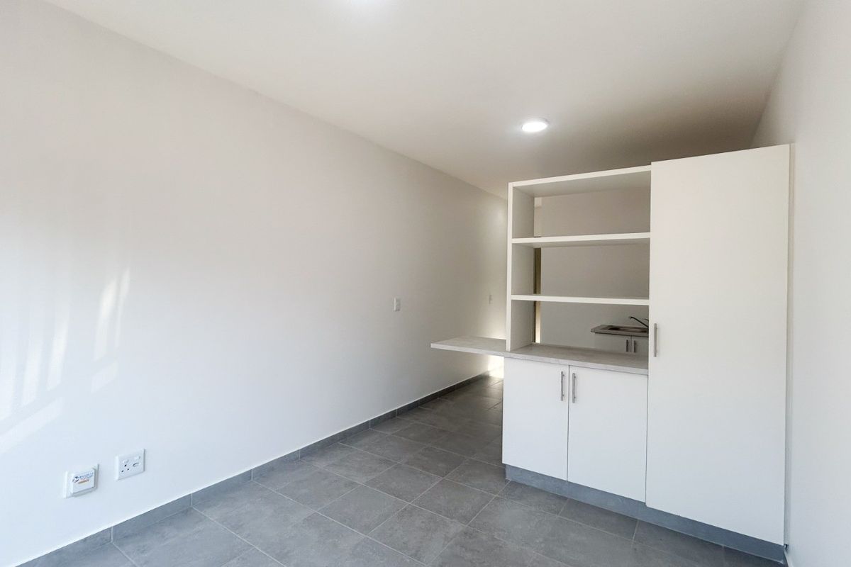 A clean, minimalist kitchen area featuring white built-in cabinetry, a small countertop, a sink, and modern grey tiled flooring.