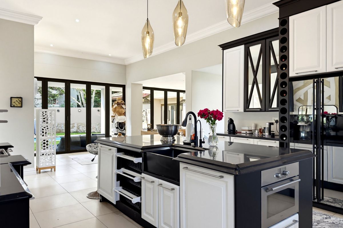 Modern kitchen featuring a large central island with a farmhouse sink, built-in oven, wine storage, and elegant pendant lighting, opening to an outdoor patio area.
