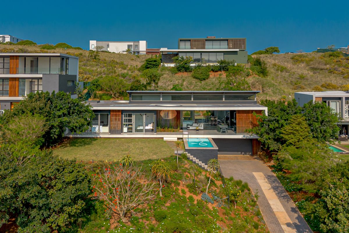 Aerial view of a modern architectural house featuring a swimming pool, paved driveway, and extensive timber and glass facade elements set in a landscaped environment.