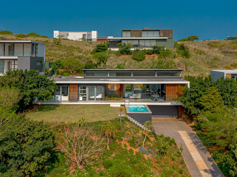 Aerial view of a modern architectural house featuring a swimming pool, paved driveway, and extensive timber and glass facade elements set in a landscaped environment.