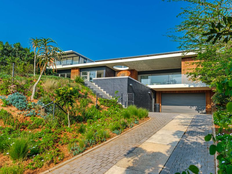 Modern multi-level residence featuring timber cladding, glass balustrades, a paved driveway, and a double garage set against a landscaped hillside.